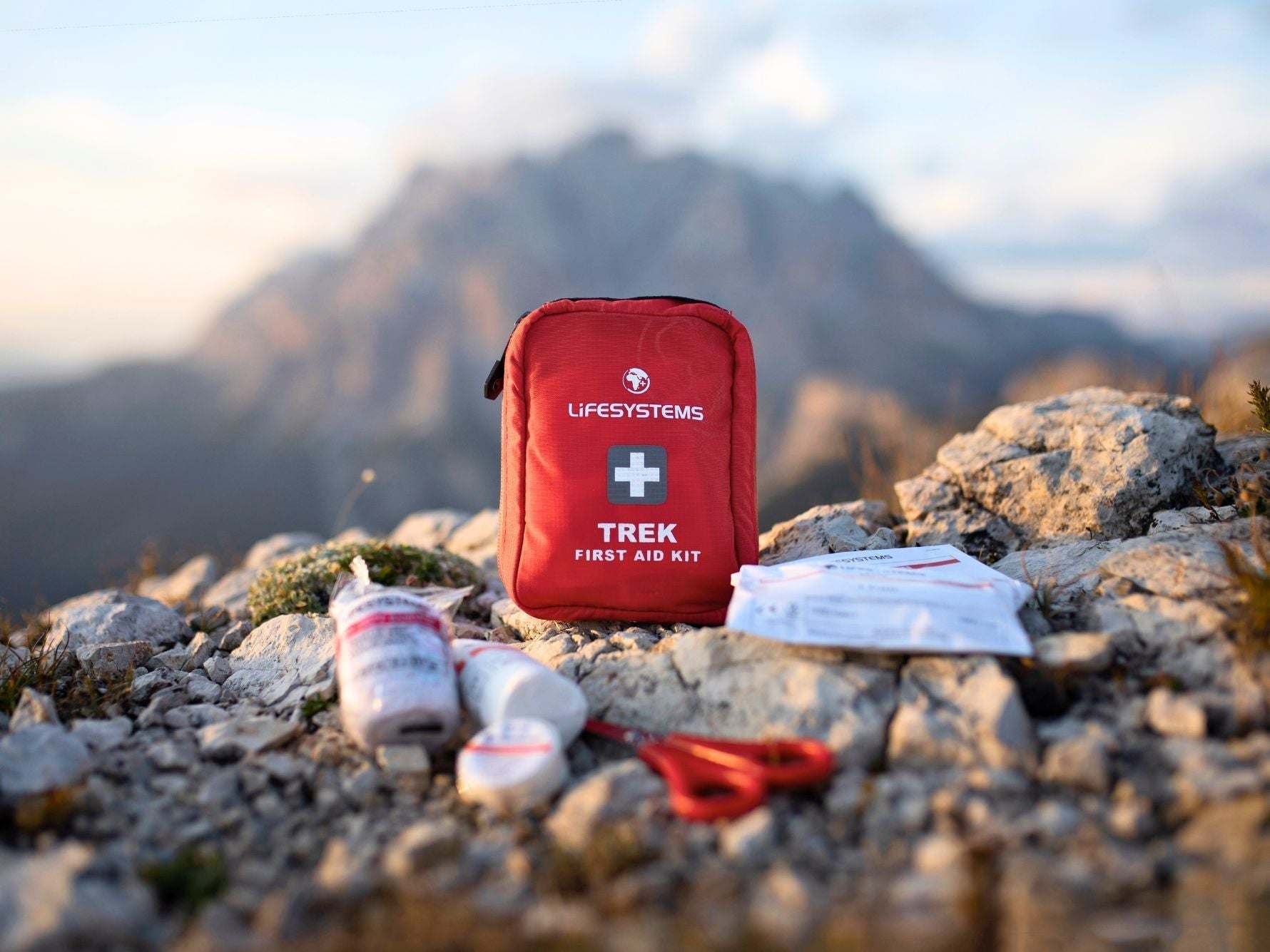 Trek First Aid Kit displayed on rocky mountain terrain with the contents laid out in front, alpine peaks in the background.