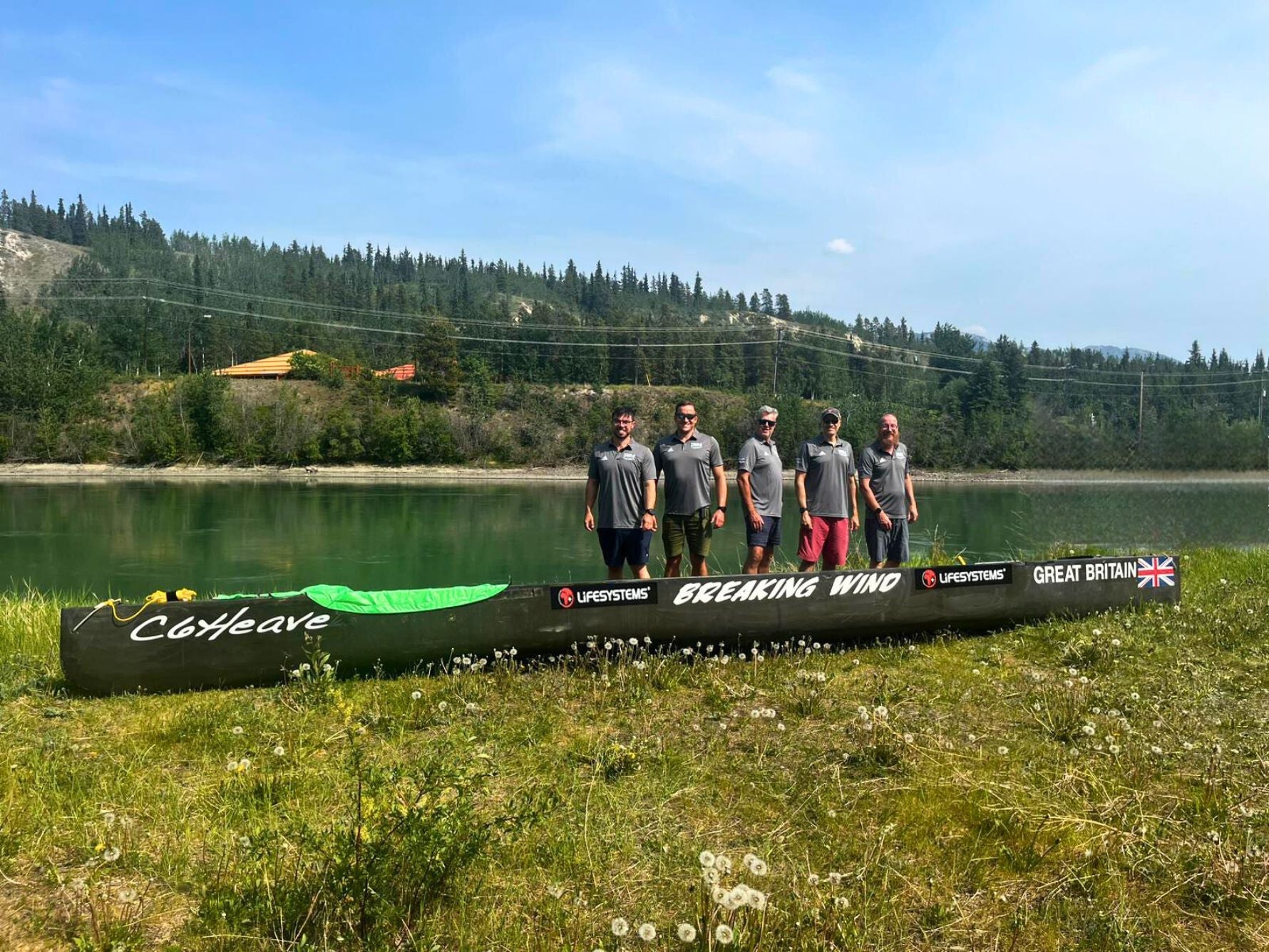 Six team members of the UK paddling crew stand beside their branded canoe 'Breaking Wind' on the riverbank ahead of the Yukon River Quest, featuring Lifesystems and Great Britain logos.