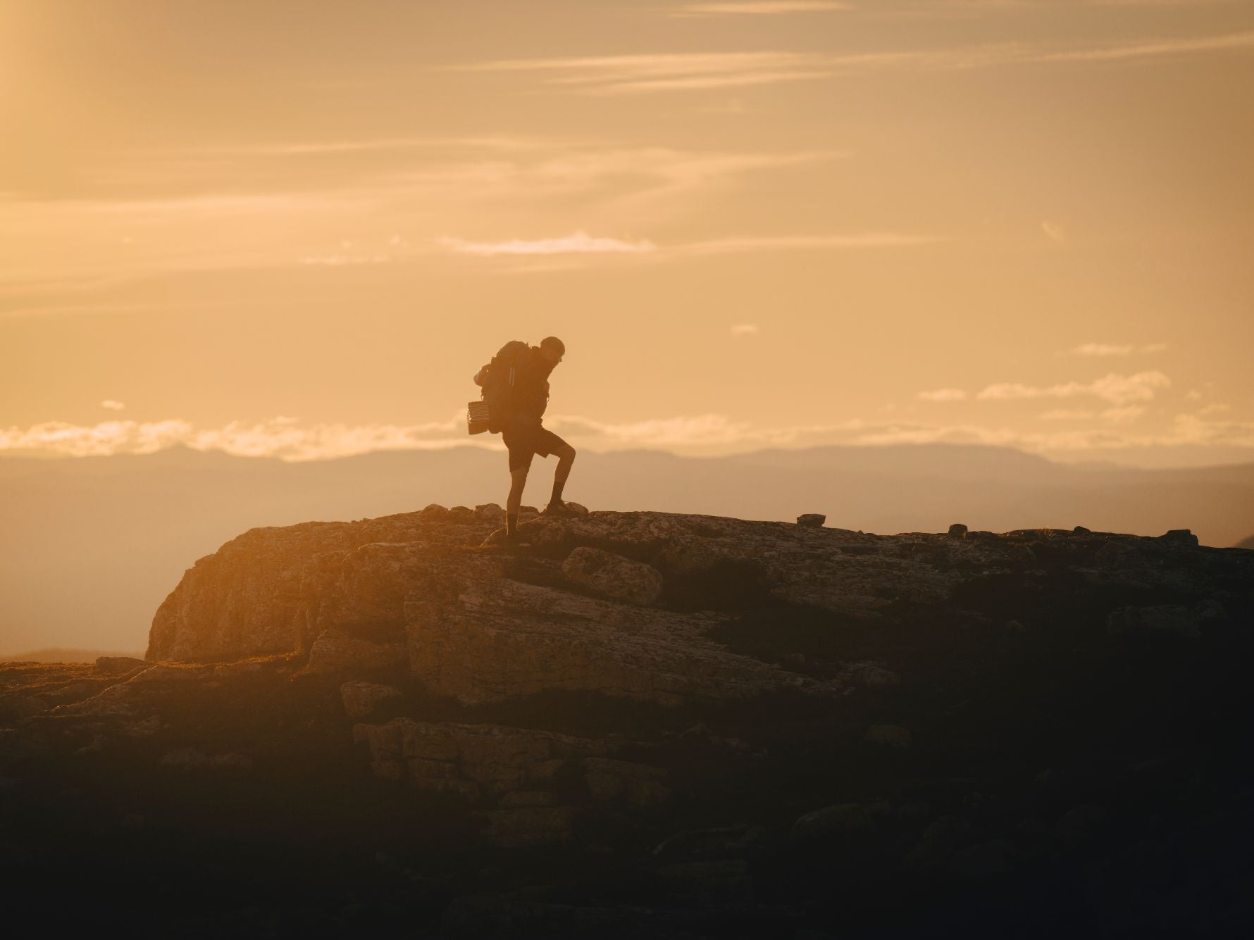 A lone hiker stands on a rocky peak at sunset, silhouetted against a golden sky with a backpack on their shoulders.