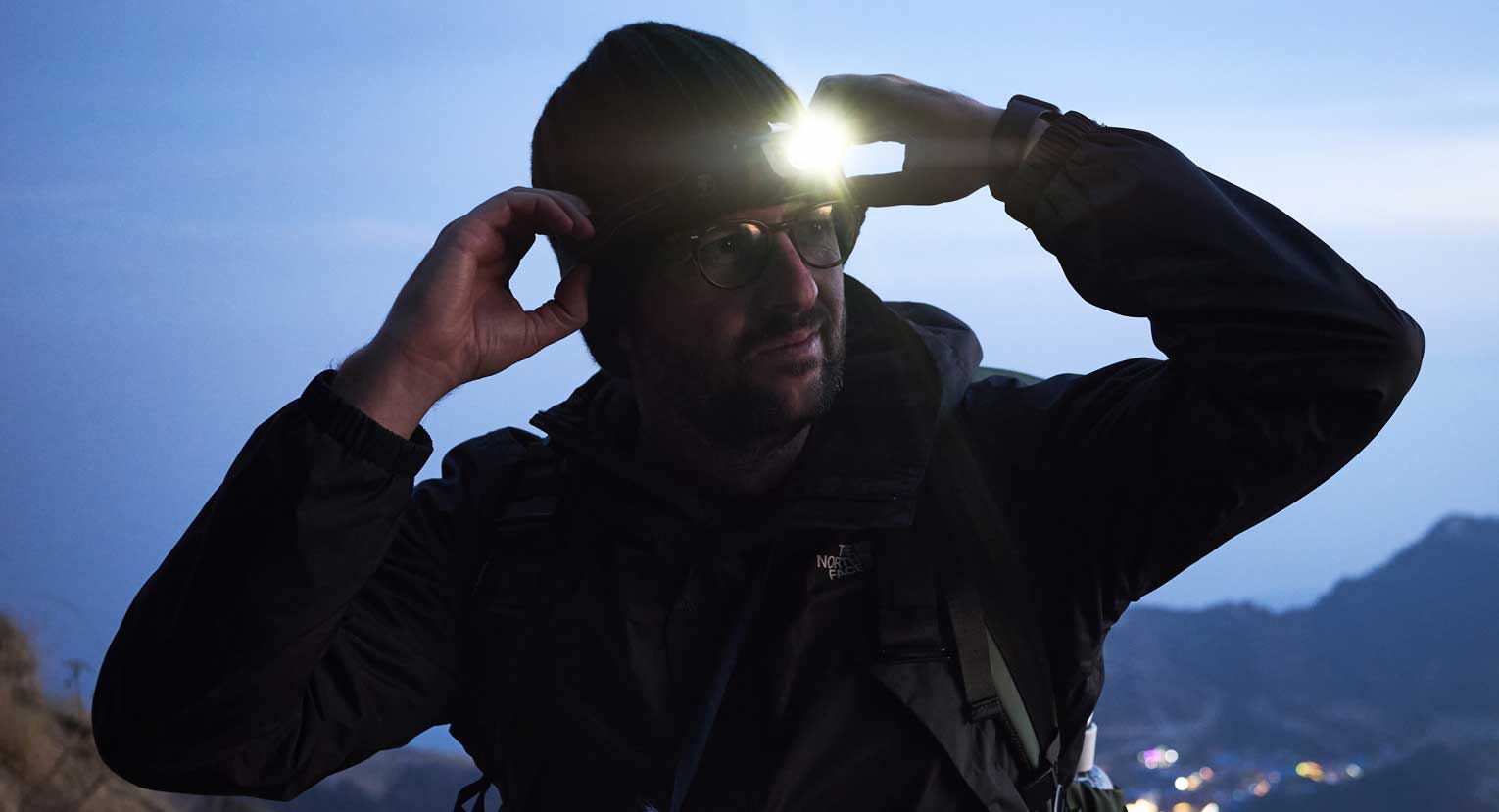 Man adjusting Lifesystems head torch on a beanie hat at dusk, standing outdoors with a mountain landscape in the background and city lights glowing below.