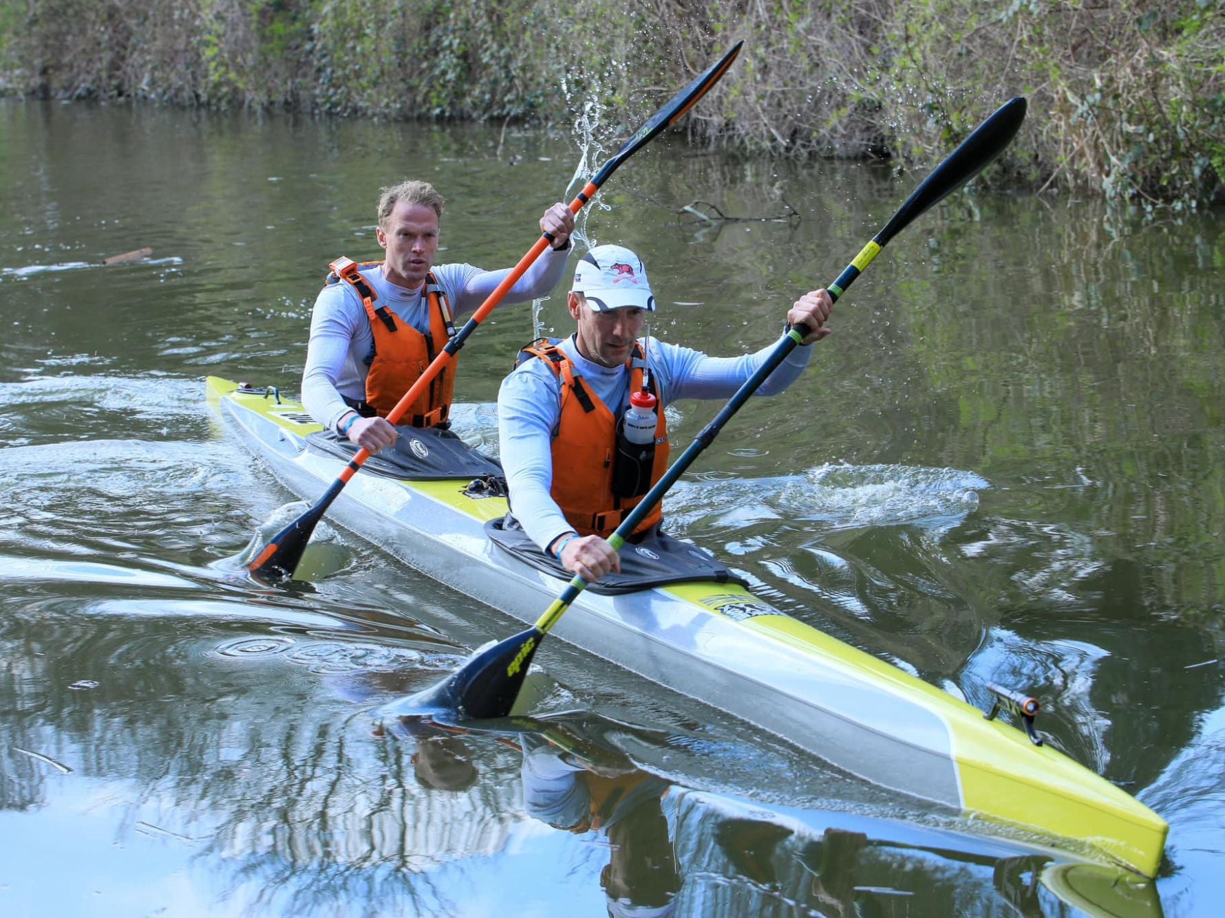 Dan Marett and Laurence Plant paddling in sync in a tandem kayak along a narrow, tree-lined waterway, both wearing matching orange life vests and focused expressions.