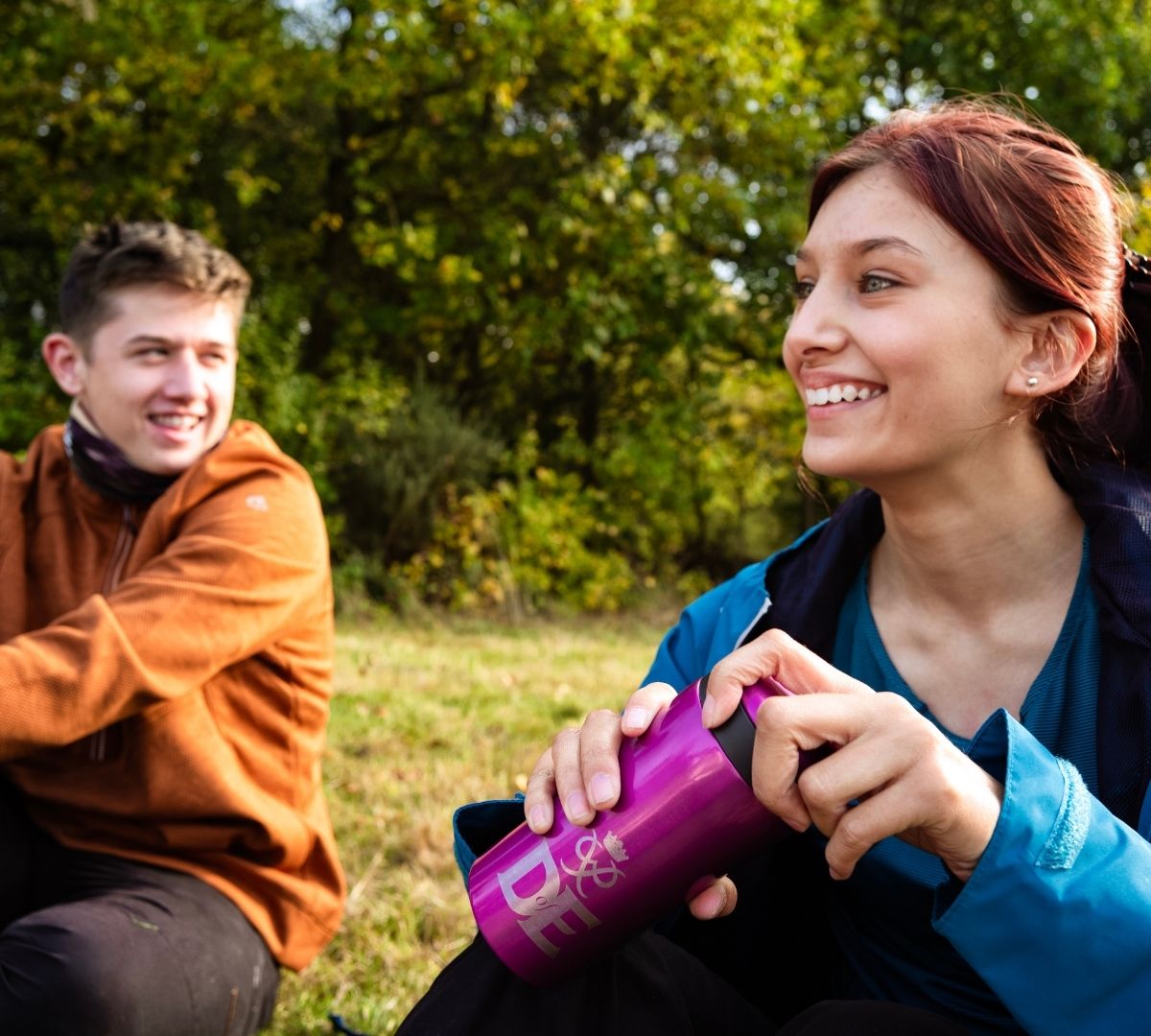 A young woman smiling while holding a bright pink Lifeventure Thermal Mug with DofE branding, sitting outdoors with a friend in a grassy, tree-lined setting.