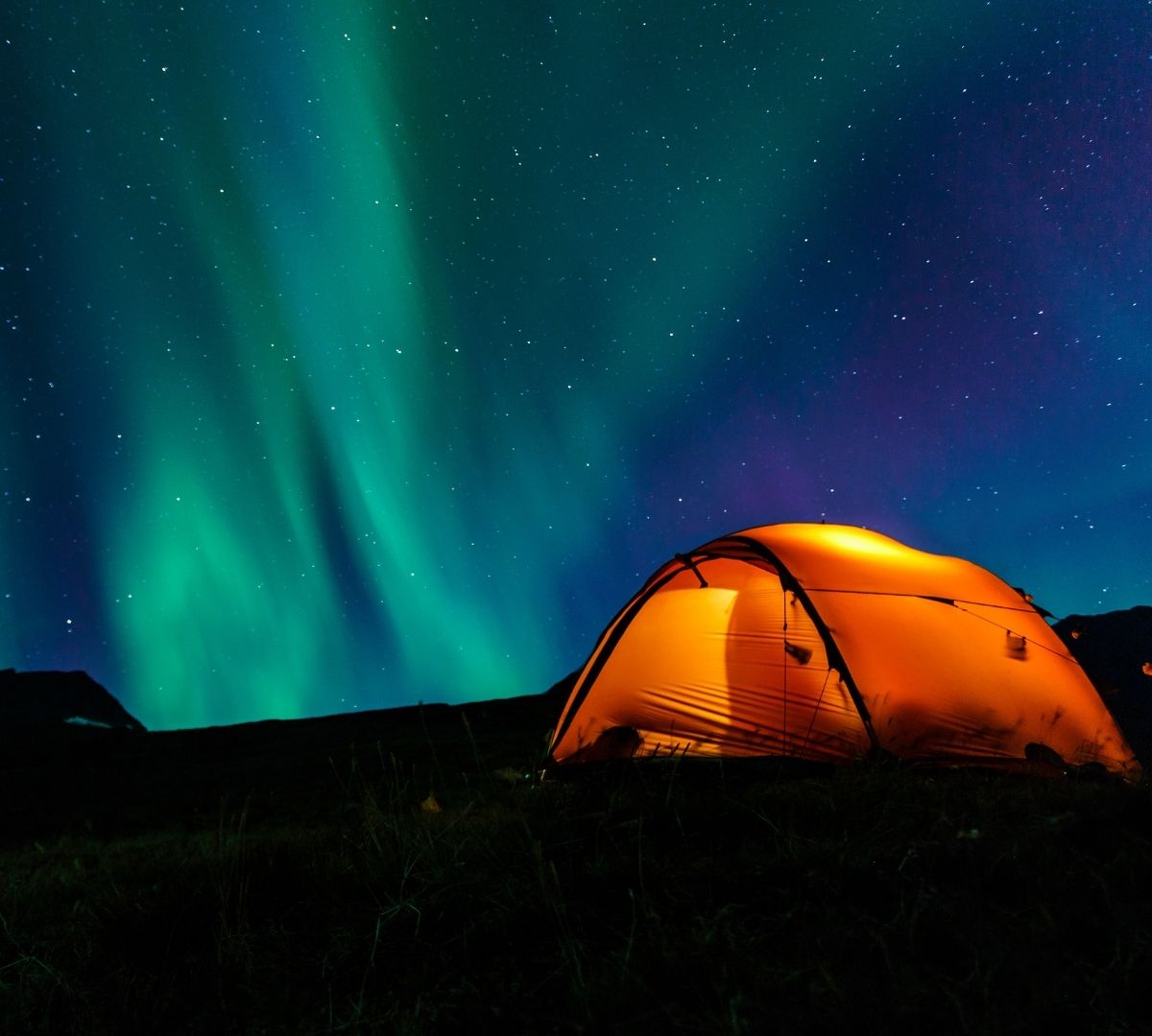 An illuminated orange tent set up in a dark landscape under a vivid green aurora borealis and starry night sky.