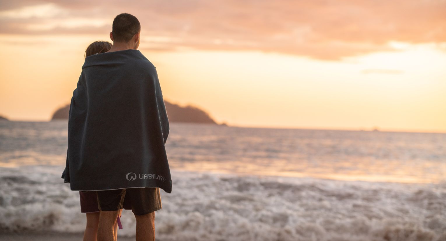 Two people wrapped in a Lifeventure towel watching the sunset by the sea.
