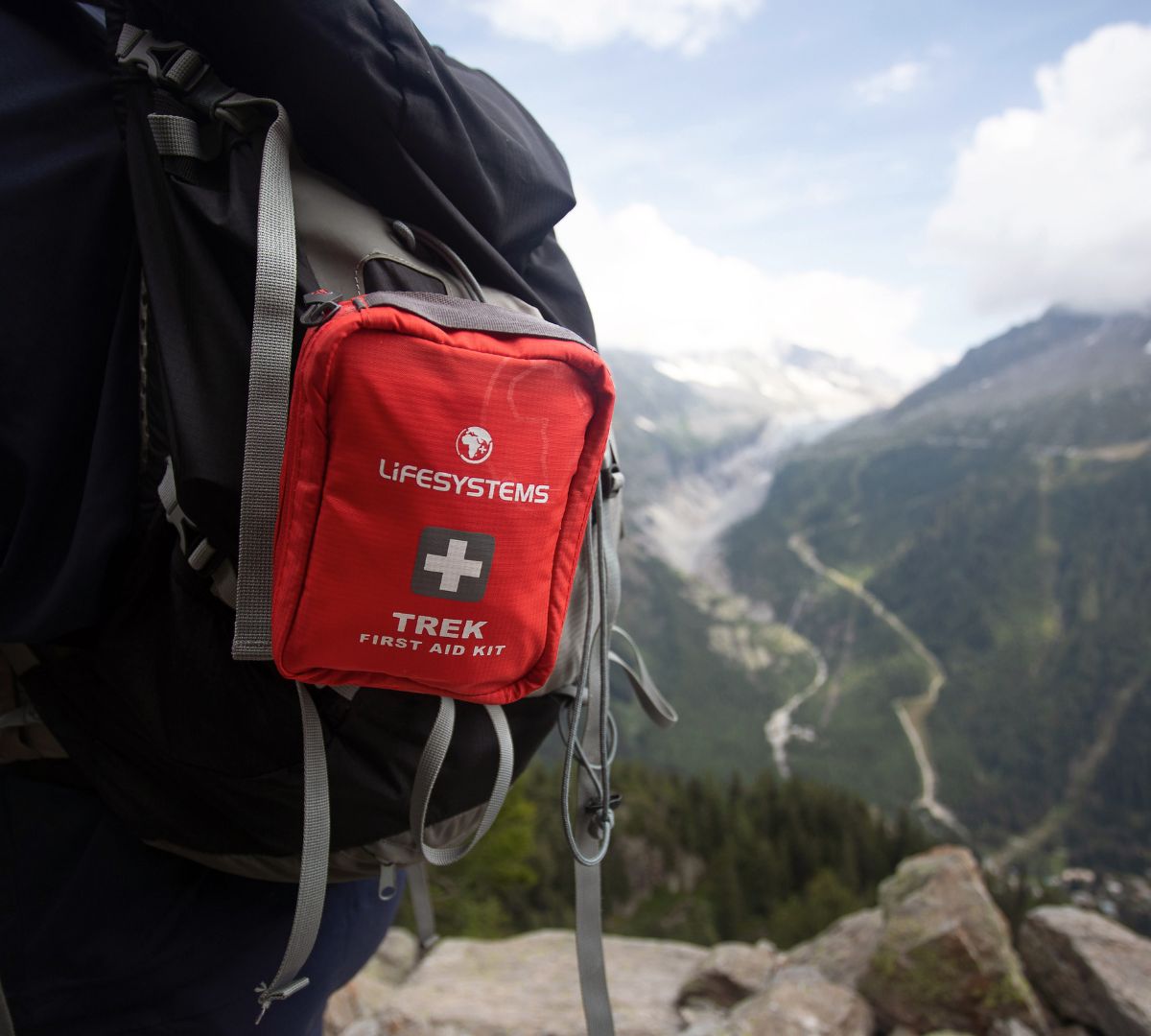 A red Lifesystems Trek First Aid Kit attached to a hiker’s backpack, overlooking a dramatic alpine valley and mountain range.