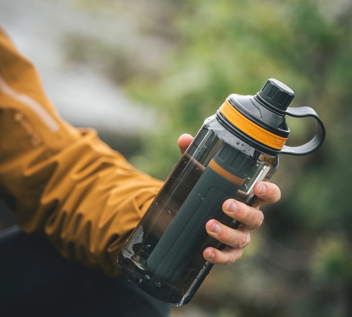 A person wearing a mustard-coloured jacket holding a Lifesystems water purifier bottle, outdoors in a lush green setting.